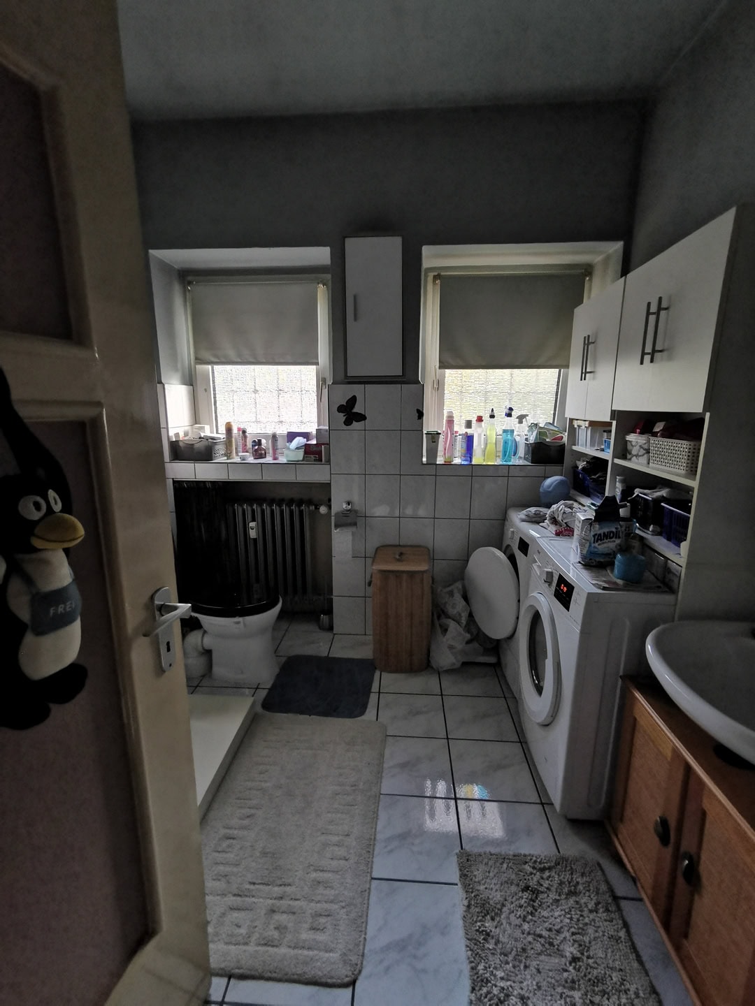 Просторная ванная комната с стиральной машиной, раковиной и окнами с шторками, много хранения и уборочной продукции, уютное пространство для домашнего комфорта.