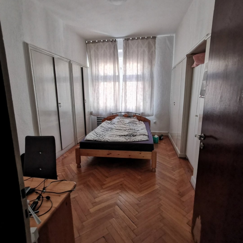 Маленькая спальня в квартире с деревянным полом и шкафами, кроватью и окнами с занавесками, интерьер в светлых тонах, уютная домашняя обстановка.