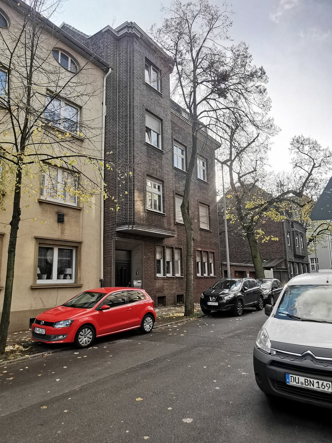 Современное жилое здание на улице с паркингом, архитектура в стиле современности, фасад с кирпичной отделкой, жилые автомобили на фоне деревьев.