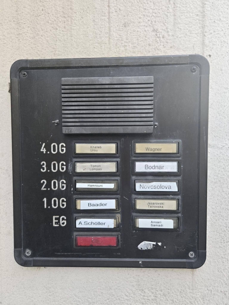 Alt: Hausnummern- und Namensschild mit verschiedenen Namen für Apartments oder Wohnungen, Außenansicht.