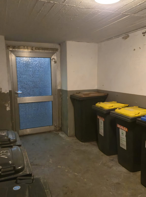 Alt: Abstellraum mit Mülltonnen und schwarzer Tür, unbequemer Raum mit Betonsockel und defekter Wand.
