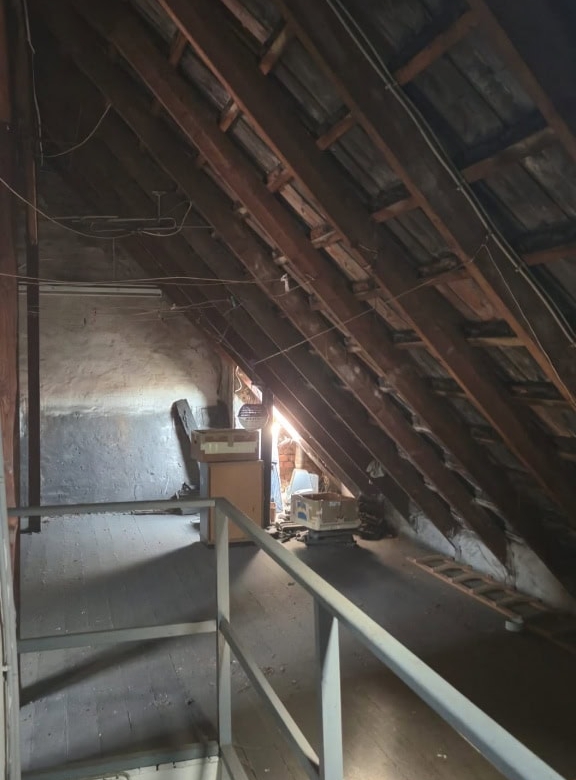 Schrägdach in einem Ausbau mit Holzbalken und Dämmmaterial, wenig natürlichem Licht, Renovierungsarbeiten, dunkler Raum, Baumaterialien und Leitungen sichtbar.