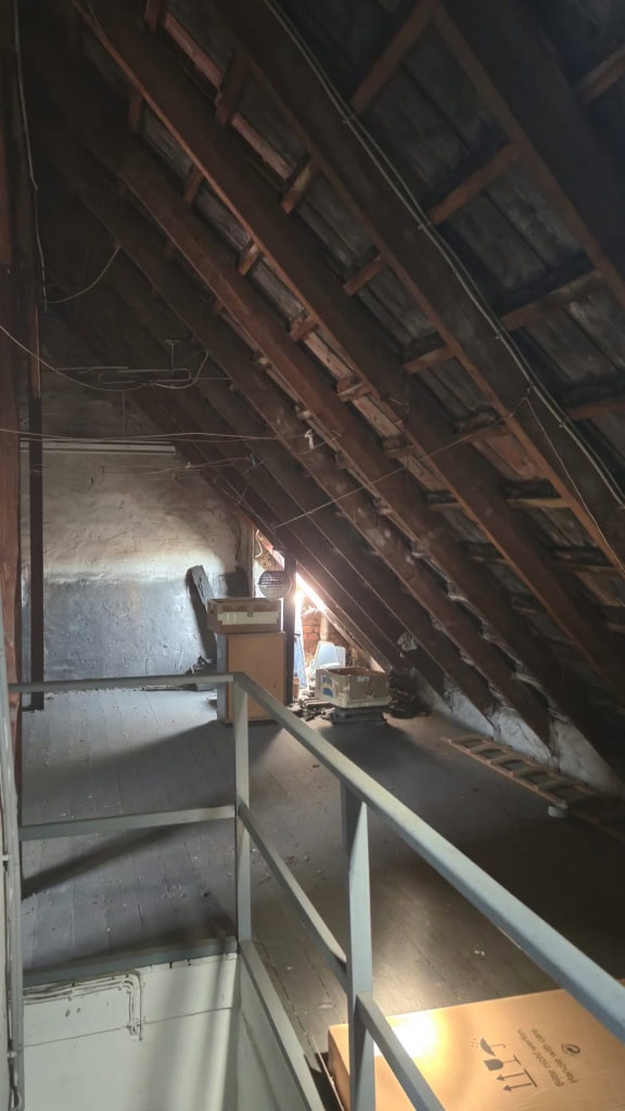 Schrägdach in einem Ausbau mit Holzbalken und Dämmmaterial, wenig natürlichem Licht, Renovierungsarbeiten, dunkler Raum, Baumaterialien und Leitungen sichtbar.